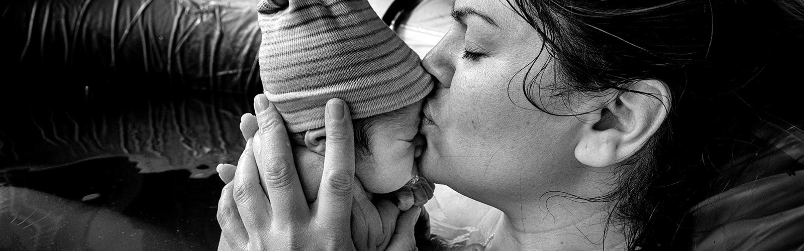 nurturing-birth-doula-directory-mobile A woman in a birthing pool holds her new born baby, her eyes are closed and she is kissing its forehead tenderly. The baby is wearing a stripy knitted hat.